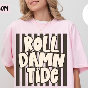 May include: A light pink short-sleeved t-shirt featuring a graphic design with vertical black stripes and the text ROLL DAMN TIDE in large, cream-colored, rounded letters centered on the front of the garment.