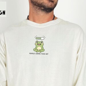 May include: An ivory long-sleeve shirt featuring a graphic of a green frog sitting in a meditative pose with a loading bar above its head and the text "MENTALLY LOADING - PLEASE WAIT" printed below the frog in a small, black, sans-serif font.