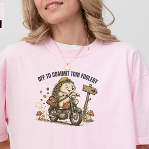 May include: A light pink t-shirt featuring a graphic of a hedgehog riding a motorcycle with a brown satchel and a yellow hat. The text above the hedgehog reads "OFF TO COMMIT TOM FOOLERY" and the shirt includes a signpost and mushrooms.