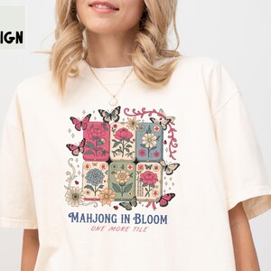 May include: An ivory short-sleeve t-shirt featuring a graphic print of six rectangular mahjong tiles decorated with various flowers and butterflies. The text "MAHJONG IN BLOOM" and "ONE MORE TILE" is printed below the tiles in blue and pink lettering.