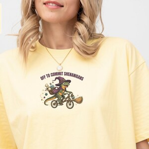 May include: A butter yellow t-shirt featuring a graphic of a dinosaur wearing a purple wizard hat and cape while riding a bicycle. The text above the dinosaur reads "OFF TO COMMIT SHENANIGANS". The shirt has short sleeves and a crew neckline.