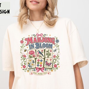 May include: An ivory t-shirt featuring a colourful graphic print with the text "MAHJONG IN BLOOM" and "ONE MORE TILE". The design includes six mahjong tiles decorated with flowers, a bird, bamboo, and a butterfly, surrounded by a floral wreath.