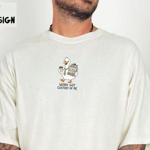 May include: An ivory t-shirt featuring a graphic of a white goose wearing a grey necktie, holding a brown coffee cup and a stack of grey file folders. Below the goose, the text reads: WORK GOT CUSTODY OF ME. The shirt has a crew neckline.