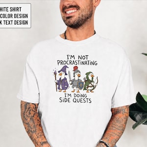 May include: A white t-shirt featuring a graphic of three ducks dressed as fantasy characters: a wizard, a knight, and an archer. The text above reads I'M NOT PROCRASTINATING and below reads I'M DOING SIDE QUESTS. The design is in full color with black text.