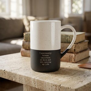 May include: A tall, two-toned ceramic mug with a speckled finish. The top half is off-white and the bottom half is black. The black section features the printed text: I accidentally became important at work and it's ruining my life. It has a curved handle.
