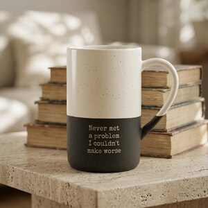 May include: A tall, two-toned ceramic mug with a speckled cream upper section and a solid black lower section. The black portion features the white printed text: Never met a problem I couldn't make worse. The mug has a curved handle on the right side.