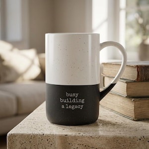 Op de afbeelding: Een tweekleurige keramische mok met een gespikkeld wit bovengedeelte en een effen zwart ondergedeelte. Het zwarte gedeelte is voorzien van de witte tekst "busy building a legacy" in een lettertype dat lijkt op een typemachine.