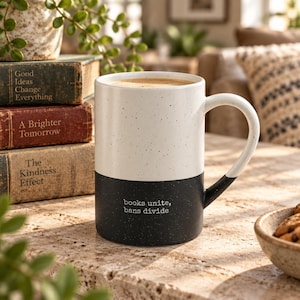 Könnte beinhalten: Ein zweifarbiger Keramikbecher mit gesprenkelter Oberfläche, bestehend aus einem weißen oberen und einem schwarzen unteren Teil. Der Text 'books unite, bans divide' ist in Weiß auf dem schwarzen Bereich aufgedruckt. Der Becher hat einen Henkel.