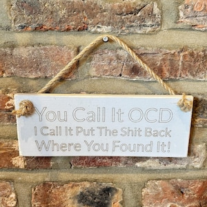 May include: White wooden sign with the text "You Call It OCD I Call It Put The Shit Back Where You Found It!" hanging on a brick wall.