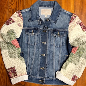 May include: A blue denim jacket with patchwork quilt sleeves. The sleeves are made of beige, white, green, and pink fabric with floral patterns. The jacket has a button closure and two front pockets.
