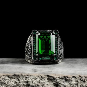 Könnte beinhalten: Ein silberner Ring mit einem großen, smaragdgrünen Edelstein, der in einem detaillierten, verzierten Design gefasst ist. Der Ring ist von kleineren, smaragdgrünen Steinen umgeben.