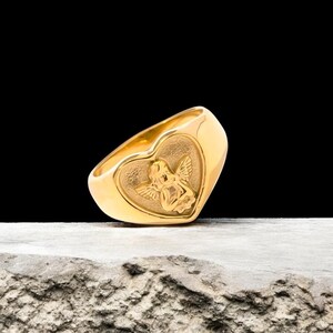 May include: A gold-colored ring with a heart-shaped design featuring an embossed cherub angel. The ring is on a light brown textured surface.