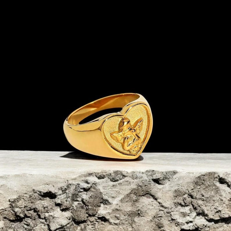 May include: A gold-toned ring with a heart-shaped design featuring a raised image of an angel. The ring is on a textured, light brown surface.