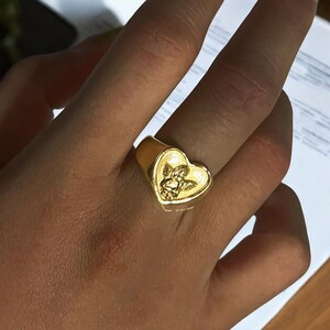 May include: A gold heart-shaped ring with an angel design. The ring is worn on a person's finger.