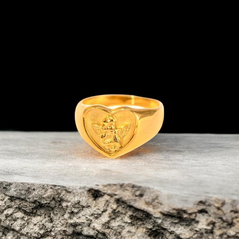 May include: A gold heart-shaped ring with an angel design. The ring is on a wooden surface.