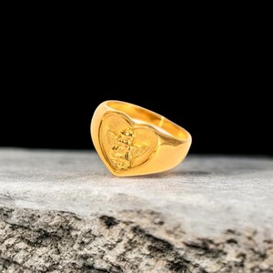 May include: A gold heart-shaped ring with an angel design. The ring is on a gray textured surface.