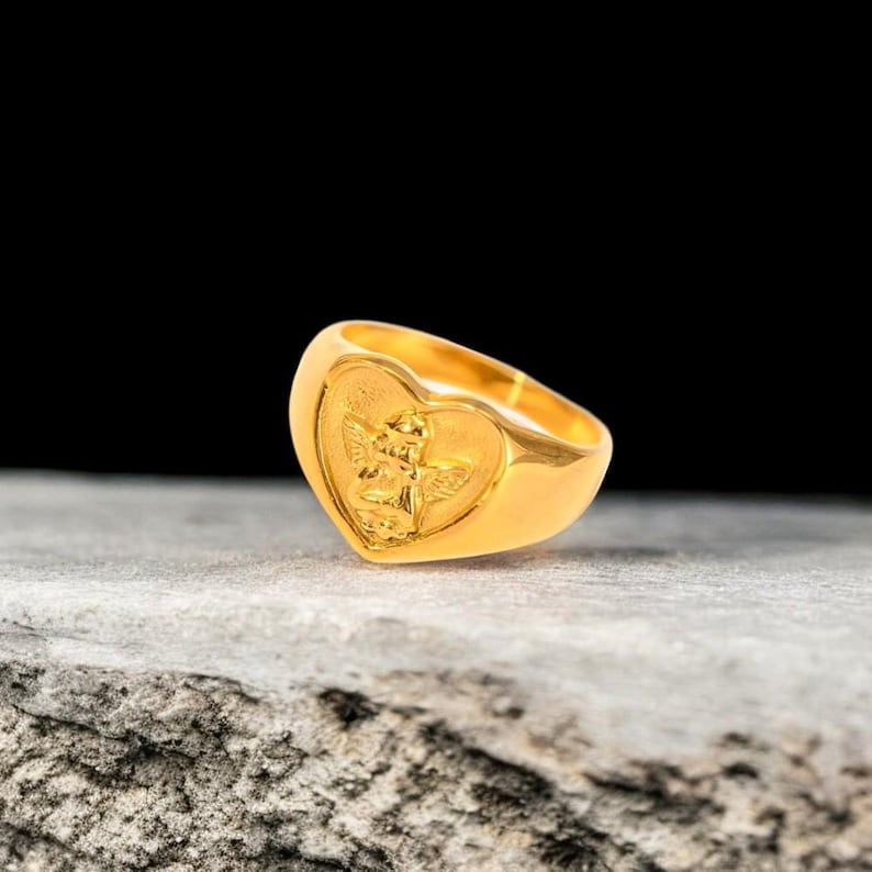 May include: A gold heart-shaped ring with an angel design. The ring is on a gray textured surface.
