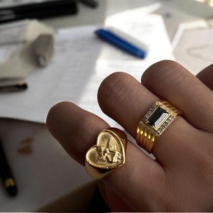 May include: Two gold rings on a hand. One ring features a heart-shaped design with an angel figure, while the other ring has a rectangular black stone with small white stones around it.