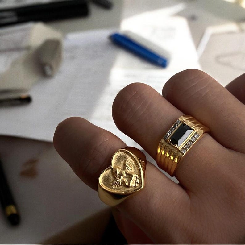 May include: Two gold rings on a hand. One ring features a heart-shaped design with an angel figure, while the other ring has a rectangular black stone with small white stones around it.