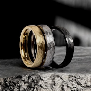 May include: Three men's wedding bands in different metals. The band on the left is gold, the middle band is silver, and the band on the right is black. All three bands have a hammered texture.