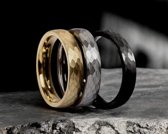 May include: Three men's wedding bands in different metals. The band on the left is gold, the middle band is silver, and the band on the right is black. All three bands have a hammered texture.