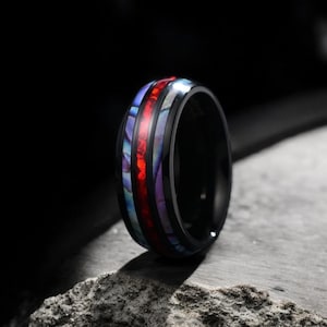 May include: A black ring with a multi-colored inlay. The inlay features red, purple, and blue colors. The ring is on a gray rock.