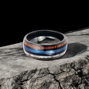 May include: A black ring with a wood grain and blue stripe design. The ring has a polished finish and is set on a gray stone surface. The ring's design includes a blue stripe, a wood grain band, and a white band.