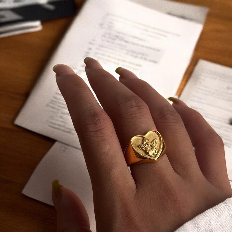 May include: A gold-tone ring with a heart-shaped design featuring an angel with wings. The ring is worn on a person's finger.