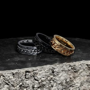 Puede incluir: Tres anillos giratorios con grabados de números romanos y un diseño de eslabón de cadena. Los anillos son negros, plateados y dorados.