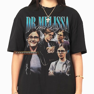 May include: Black t-shirt with a vintage-style graphic of Dr. Melissa Mel King. The design includes multiple images of the doctor, the text "DR. MELISSA" in a blue gradient, and the name "Mel King".