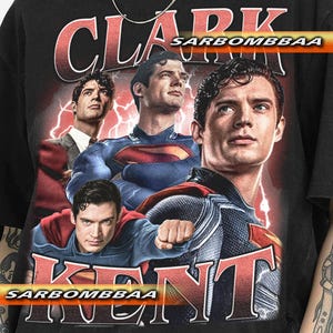May include: Black t-shirt featuring a graphic of Clark Kent in various poses, with the text "CLARK KENT" in red. The shirt has a vintage look, with a faded design and the text "SARBOMBBAA".