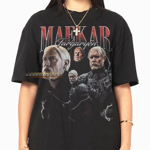 Op de afbeelding: Zwart T-shirt met een afbeelding van meerdere mannen met wit haar en baarden, met de tekst "MAEKAR Targaryen" in rood en wit. Het ontwerp heeft een vintage, verweerde uitstraling.