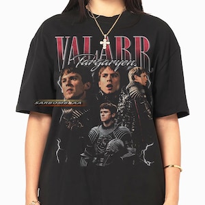 以下が含まれることがあります： 男性のグラフィックと「VALARR Targaryen」のテキストが赤と白でプリントされた黒いTシャツ。 ヴィンテージスタイルで、稲妻のアクセントと「SARBOMBBAA」のテキストが特徴です。