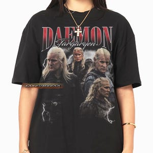 Puede incluir: Camiseta negra con un gráfico de un personaje con pelo largo y rubio y el texto "DAEMON Targaryen" en rojo y blanco. La camiseta tiene un diseño vintage y desgastado, con múltiples imágenes del personaje. La marca "SARBOMBBAA" está impresa en la camiseta.