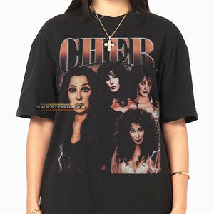 Może przedstawiać: Czarny t-shirt z grafiką Cher i napisem "CHER" w pomarańczowych i białych literach. Projekt zawiera wiele portretów Cher. Koszulka ma okrągły dekolt.