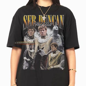 May include: Black t-shirt with a retro design featuring Ser Duncan the Tall from a fantasy series. The graphic includes images of the character in armour and the text "SER DUNCAN the Tall" in gold. The brand name "SARBOMBBAA" is also visible.