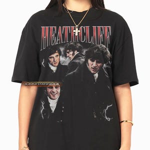 Puede incluir: Camiseta negra con el nombre "HEATHCLIFF" en letras rojas y blancas. La camiseta presenta un collage de imágenes de un hombre. El diseño tiene un aspecto vintage y desgastado.