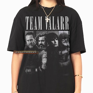 以下が含まれることがあります： 「TEAM VALARR」の文字が白でプリントされた黒いTシャツ。4人の男性の白黒画像が上にあります。Tシャツは半袖で、クルーネックです。
