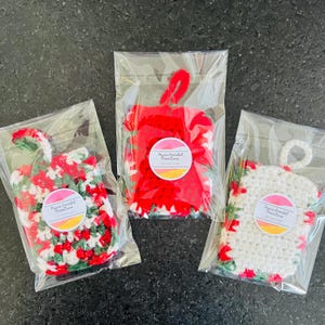 Könnte beinhalten: Drei gehäkelte Ornamente in einzelnen Plastiktüten. Die Ornamente sind rot, weiß und grün mit einem weißen runden Etikett, auf dem "Maya Crochet Creations" steht.