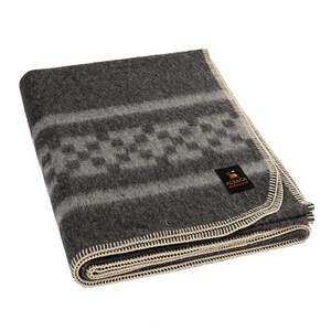 Peut inclure: Une couverture en laine d'alpaga grise avec un motif géométrique blanc et gris. La couverture a une bordure crème et une étiquette qui indique "ALPACCA WAREHOUSE".