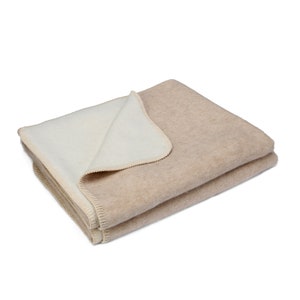 May include: A beige and white reversible blanket made of wool. The blanket is folded in half and the beige side is facing up.