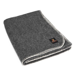 Peut inclure: Une couverture en laine d'alpaga de couleur gris anthracite avec une bordure blanche cousue. La couverture est pliée et porte une petite étiquette sur laquelle est inscrit "Alpaca Wool Products" en blanc sur fond noir.