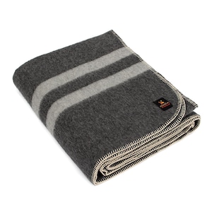 Peut inclure: Une couverture en laine grise avec deux bandes blanches. La couverture est pliée et a une bordure cousue de couleur crème. La couverture porte une petite étiquette avec le texte "Faribault Woolen Mill".