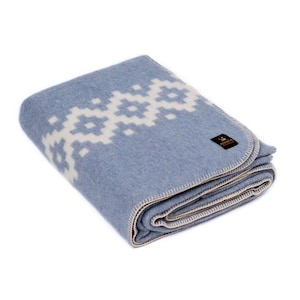 May include: A folded blue wool blanket with a white geometric pattern. The blanket has a cream-colored border and a small label with the word "ALASKA" on it.