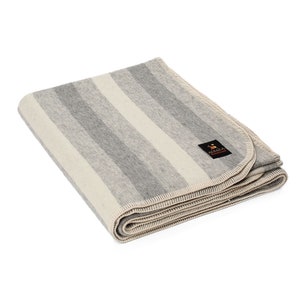 Peut inclure: Une couverture en laine d'alpaga à rayures grises et blanches avec une étiquette brune indiquant "Alpaca" et un petit logo d'alpaga. La couverture est pliée et a une bordure blanche.