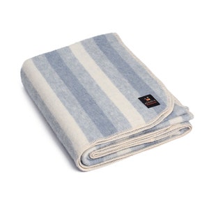 Peut inclure: Une couverture en laine d'alpaga rayée bleu et blanc avec une bordure crème. La couverture est pliée et le logo "Alpaca Designs" est visible dans le coin inférieur droit.
