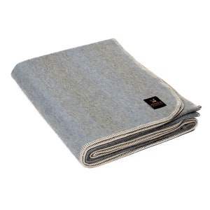 Peut inclure: Une couverture pliée, de couleur gris clair, avec une bordure cousue de couleur crème. La couverture comporte une petite étiquette noire avec du texte. Les tons doux et neutres de la couverture suggèrent un textile de maison confortable.