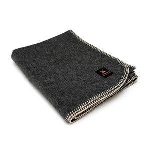 May include: A folded charcoal gray blanket with a cream-colored stitched border. The blanket has a small black label with the word "Alpaca" and a logo. The blanket is made of a soft, textured material.