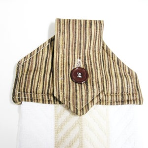 May include: A hanging kitchen towel featuring a white fabric base with a subtle herringbone pattern. The top section is made of brown and tan striped fabric, folded over with a pointed tab secured by a single dark brown plastic button.