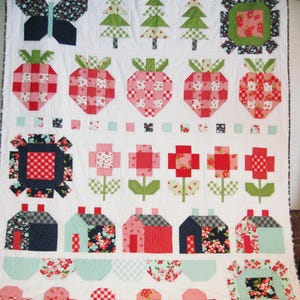 May include: A white quilt featuring a patchwork design with various motifs including a dark butterfly, three green pine trees, red strawberries, floral squares, red flowers with green stems, and five small houses in red, blue, and gray.